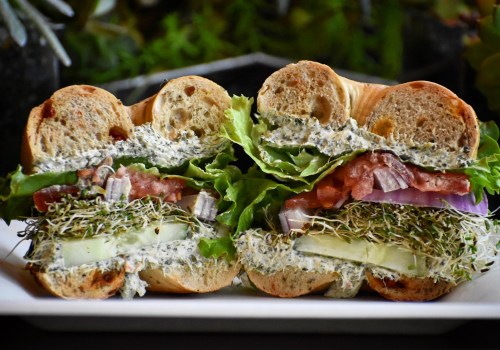 A close-up of a sandwich on a plate, featuring layers of lettuce, tomato, cucumber, and a creamy spread between bagel halves.