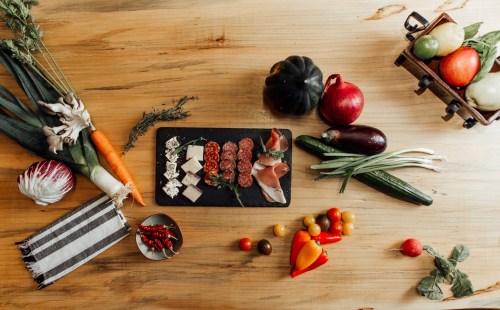 A wooden table with various fresh vegetables and a platter of meats and cheeses arranged on a slate board.