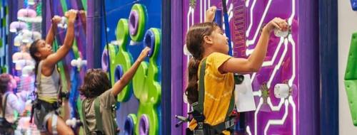 Children climbing colorful indoor rock walls, focused and wearing safety harnesses.