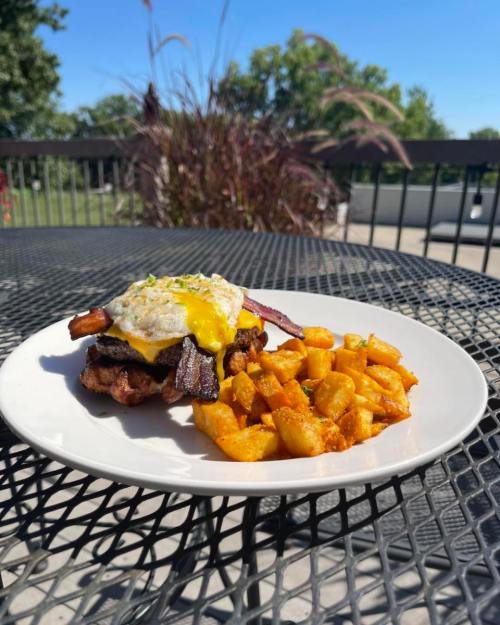 A plate of crispy potatoes and a sandwich topped with bacon, a fried egg, and a sunny yellow sauce, outdoors.
