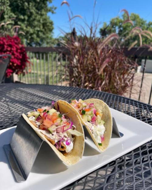 Two tacos in a metal holder, filled with colorful toppings, on a white plate with a sunny outdoor background.