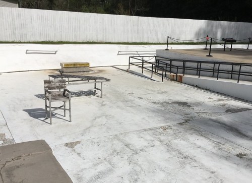 An empty outdoor space with a white concrete floor, featuring a table and chair, and a raised platform in the background.