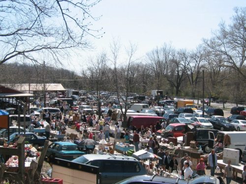 A bustling outdoor flea market with numerous stalls, cars, and people browsing under a clear blue sky.