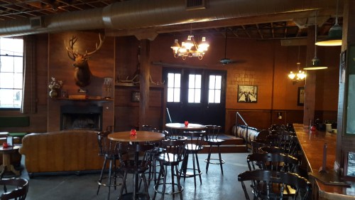 Cozy interior of a rustic bar with wooden furniture, a fireplace, and a mounted deer head on the wall.