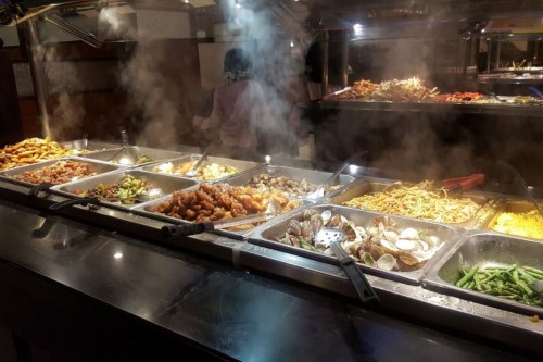 A buffet with various dishes, including noodles, vegetables, and seafood, surrounded by steam and warm lighting.