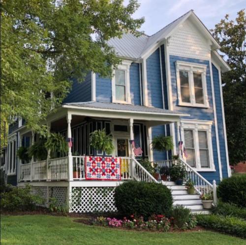 A charming blue house with a white porch, decorated with plants and a colorful quilt, surrounded by a lush garden.