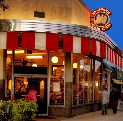 A charming restaurant with red and white awnings, glowing lights, and people walking by in the evening.