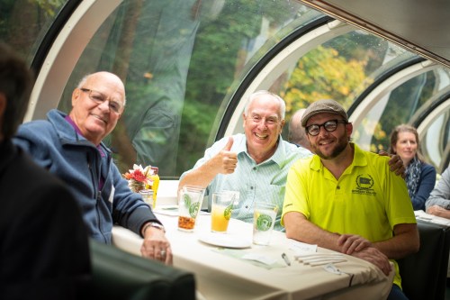 Three men smiling at a table in a train dining car, with drinks and a scenic view through large windows.