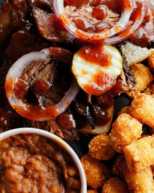 A close-up of a BBQ plate featuring sliced brisket with sauce, pickles, onions, baked beans, and crispy tater tots.
