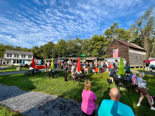 A lively outdoor event with people gathered near a barn, surrounded by tables and colorful umbrellas under a blue sky.