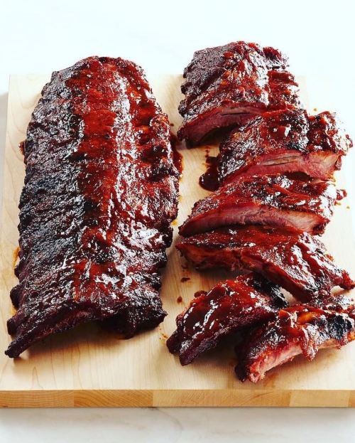 A wooden cutting board with a large rack of glazed ribs and several sliced pieces, showcasing a rich, smoky appearance.