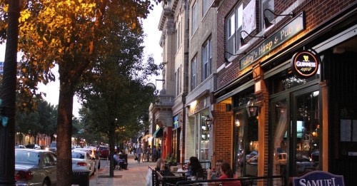 A lively street scene with shops and restaurants, featuring outdoor seating and trees illuminated by evening light.