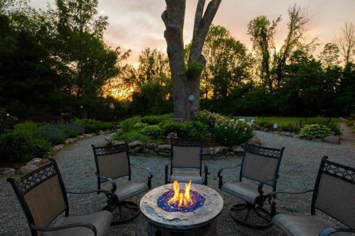 A cozy outdoor fire pit surrounded by chairs, with a sunset and trees in the background.