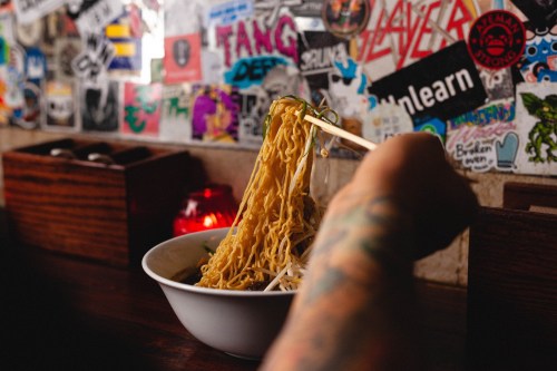 A hand holding chopsticks lifts noodles from a bowl, with a colorful, sticker-covered wall in the background.