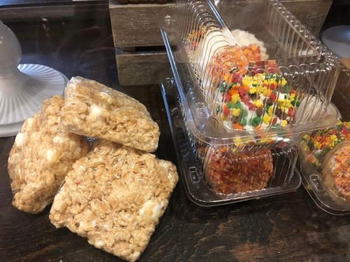A stack of rice crispy treats next to a clear container filled with colorful frosted desserts.