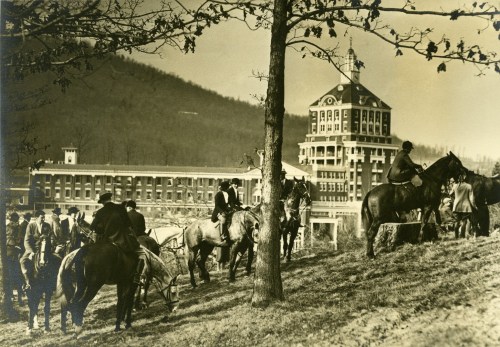 Historic black-and-white photo of people on horseback near a large building, with trees and hills in the background.
