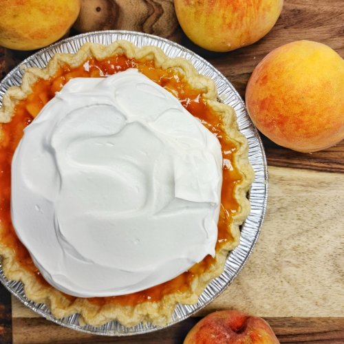 A peach pie topped with whipped cream, surrounded by fresh peaches on a wooden surface.