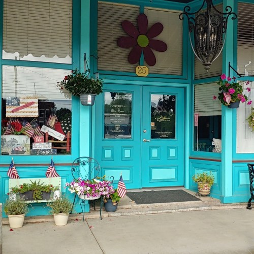 Bright turquoise storefront with a large flower decoration, hanging plants, and American flags adorning the entrance.