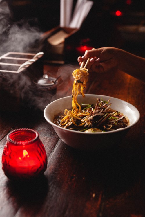 A hand using chopsticks to lift noodles from a steaming bowl, with a red candle glowing nearby.