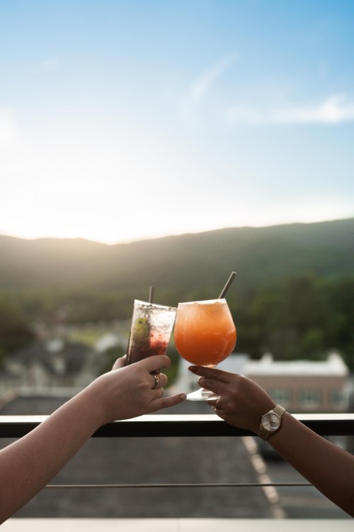 Two hands clink colorful cocktails against a scenic sunset backdrop with mountains in the distance.