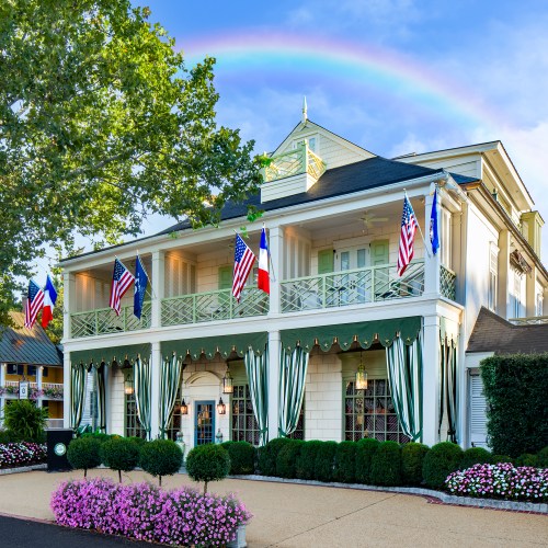A charming building with flags, green awnings, and colorful flowers, under a rainbow in a clear blue sky.