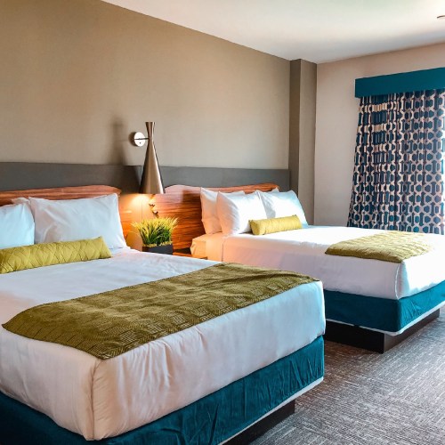A modern hotel room featuring two beds with green accents, a window with patterned curtains, and a cozy atmosphere.