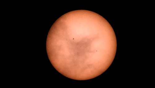 A close-up view of the sun, partially obscured by clouds, with dark spots visible on its surface.