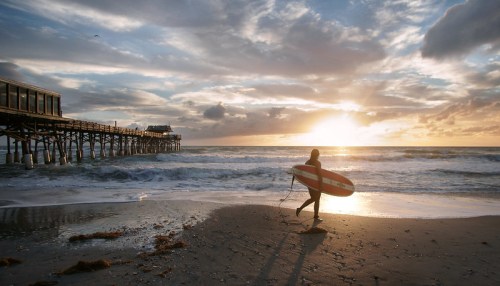 A surfer walks along the beach at sunset, with waves crashing and a pier in the background under a colorful sky.