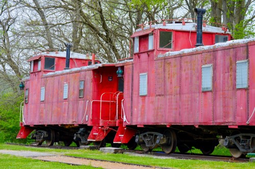 Two vintage red cabooses sit on tracks surrounded by greenery and trees, showcasing weathered paint and classic design.