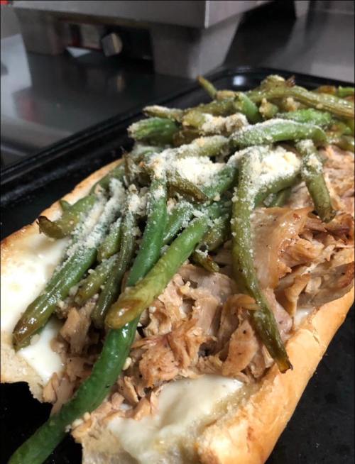 A sandwich with shredded meat, melted cheese, and topped with green beans and grated cheese on a toasted hoagie roll.