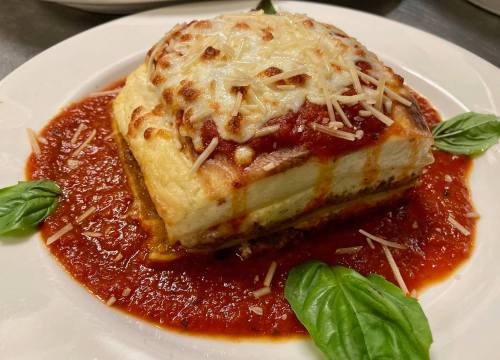 A square slice of lasagna topped with melted cheese, served on a bed of marinara sauce and garnished with basil leaves.