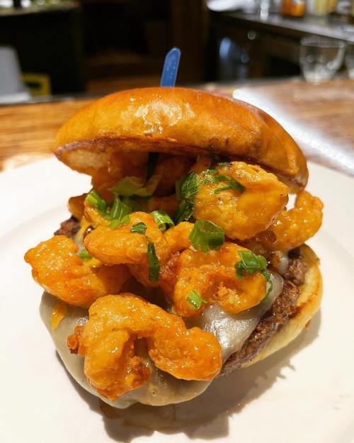 A close-up of a burger topped with crispy shrimp, melted cheese, and green onions on a toasted bun.