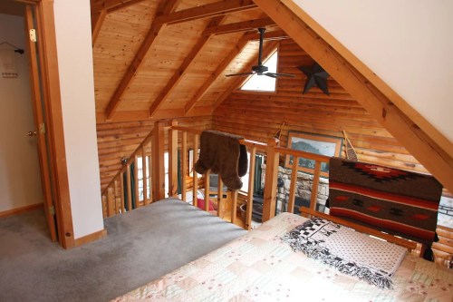 Cozy loft space with wooden beams, a quilted bed, and a view of a rustic living area below.