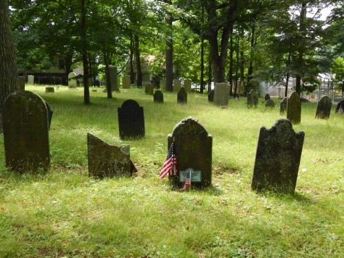 A peaceful cemetery with old gravestones surrounded by trees and grass, featuring small American flags at some graves.