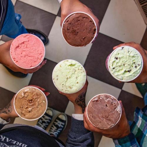 Six hands hold ice cream cups in various flavors, arranged in a circle on a checkered floor.