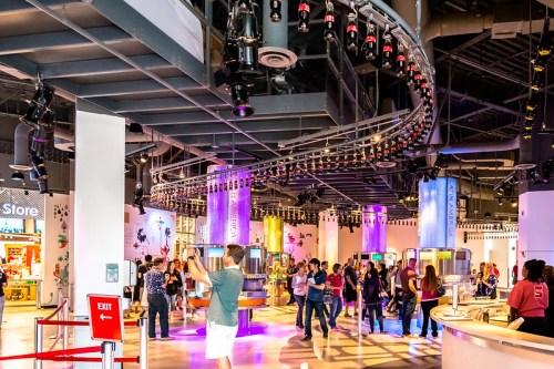 A vibrant indoor space with people exploring, colorful lights, and a roller coaster track overhead.