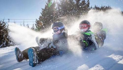 Three children sledding down a snowy hill, surrounded by snow spray and bright sunlight.