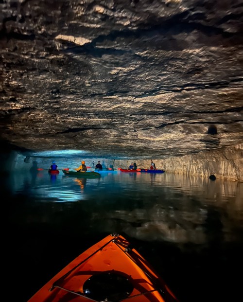 A group of kayakers paddles through a dark cave with a glowing blue light reflecting on the water's surface.