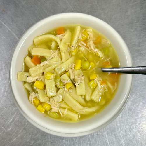 A bowl of chicken noodle soup with vegetables, including corn, peas, and carrots, on a metal surface.