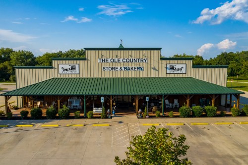 A large country store and bakery with a green roof, featuring a porch and outdoor seating, set against a blue sky.
