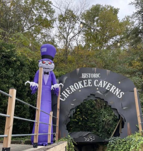 A large purple inflatable figure stands near the entrance of Cherokee Caverns, surrounded by trees and autumn foliage.