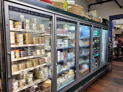 A refrigerated display case filled with various jars, containers, and packaged foods in a market setting.