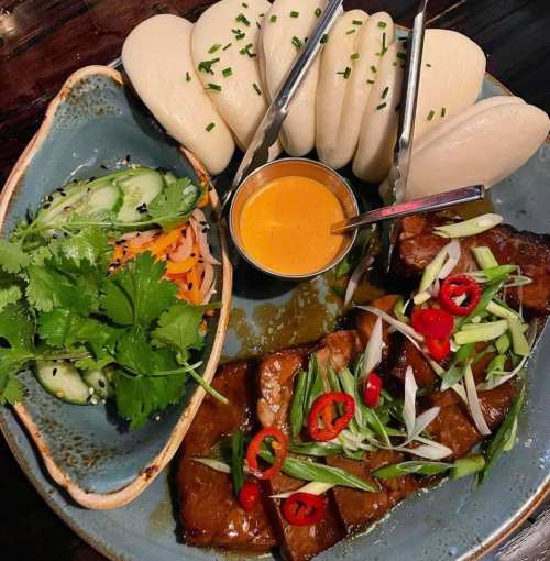 A plate of grilled meat with bao buns, cucumber salad, and a dipping sauce, garnished with herbs and chili slices.