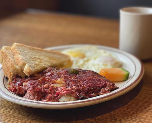 A plate with corned beef hash, two eggs, toast, and a slice of melon, with a coffee cup in the background.