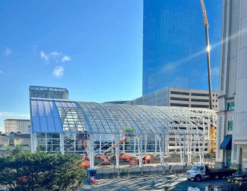 Construction site featuring a large glass structure with cranes and machinery, set against a blue sky and tall buildings.