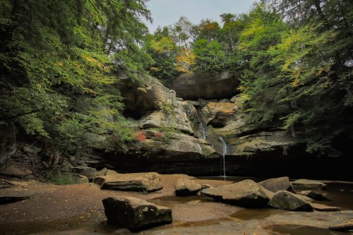 A serene landscape featuring a rocky cliff with a waterfall, surrounded by lush green trees and a sandy area below.