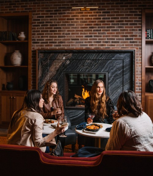 Four women sit around a table with food and drinks, chatting in a cozy setting by a fireplace.