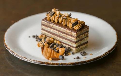 A layered chocolate dessert topped with nuts and drizzled caramel on a white plate.