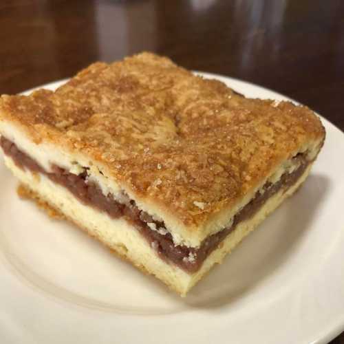 A slice of layered dessert with a golden, crumbly top and a chocolate filling, served on a white plate.