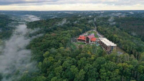 Aerial view of a lodge surrounded by lush green forests and misty hills under a cloudy sky.